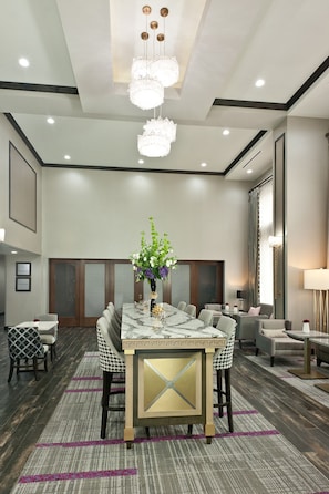 A modern hotel room with a marble dining table, comfortable seating, a chandelier, and a large window with curtains.