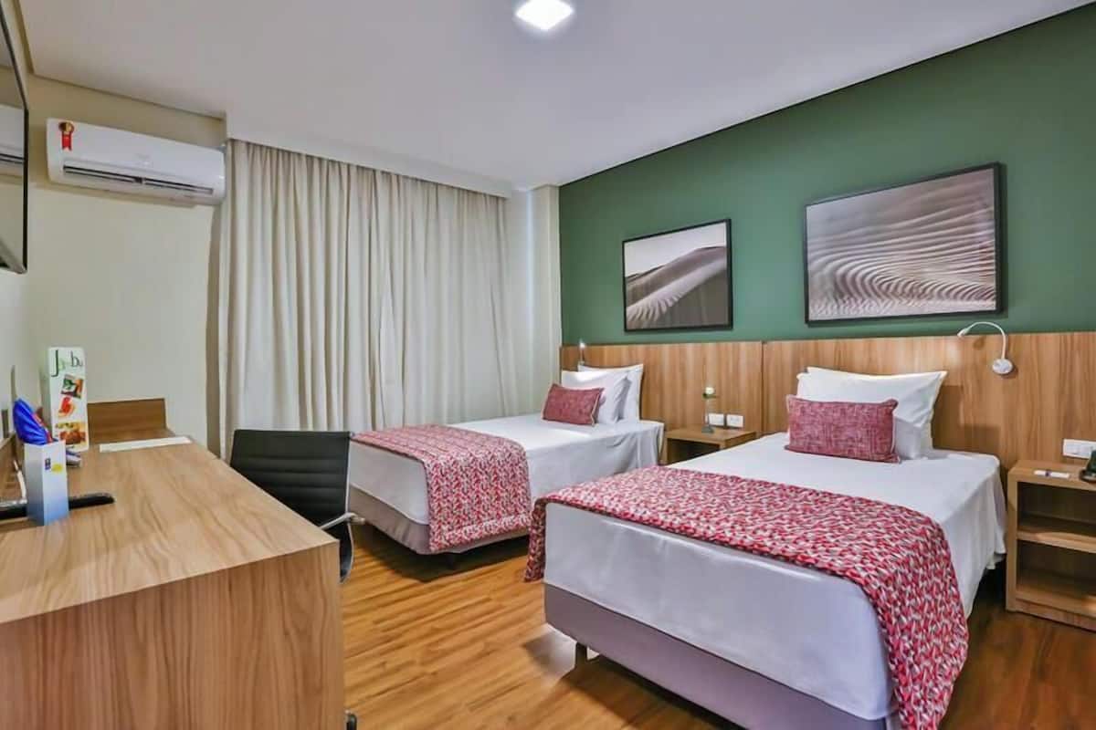 A hotel room with two beds, a desk, and a chair. There is an air conditioning unit on the wall, a window with curtains, and two paintings on the wall.