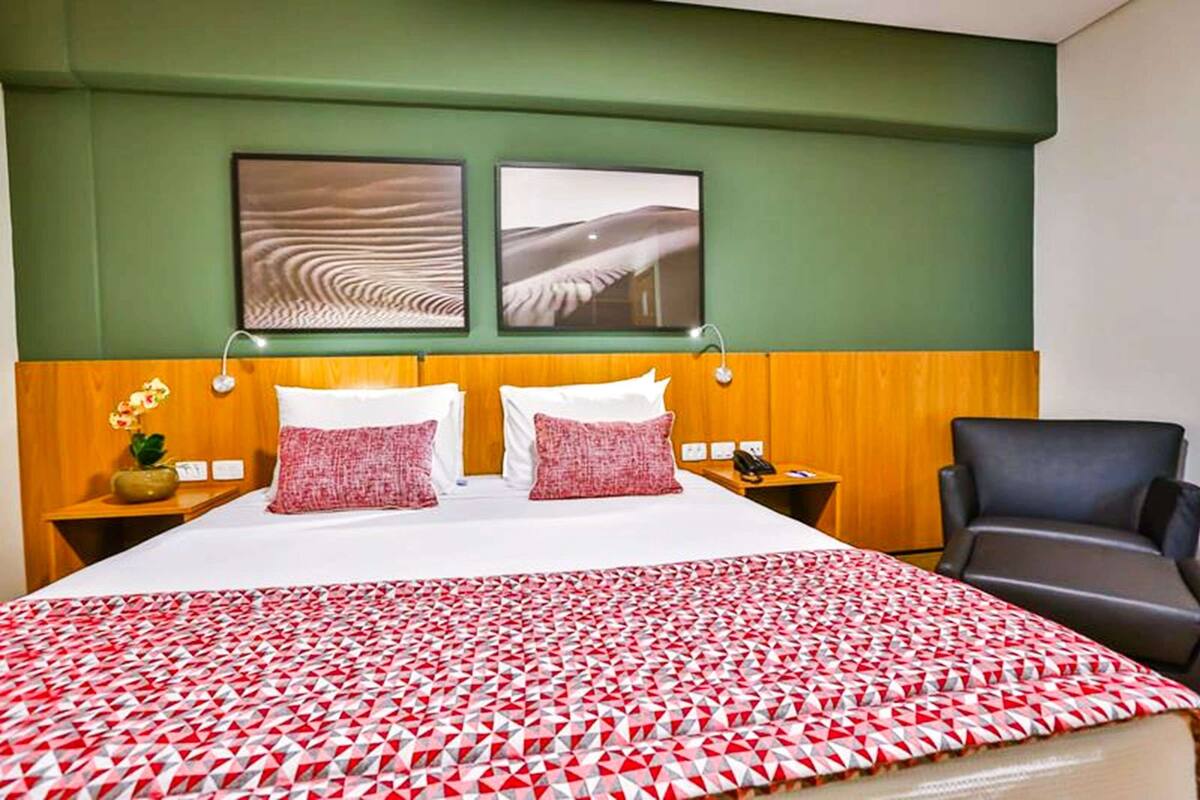 A hotel room with a large bed, two pillows, a red and white patterned bedspread, a wooden headboard, two wall-mounted artworks, a nightstand with a phone, a small plant, and a black armchair.