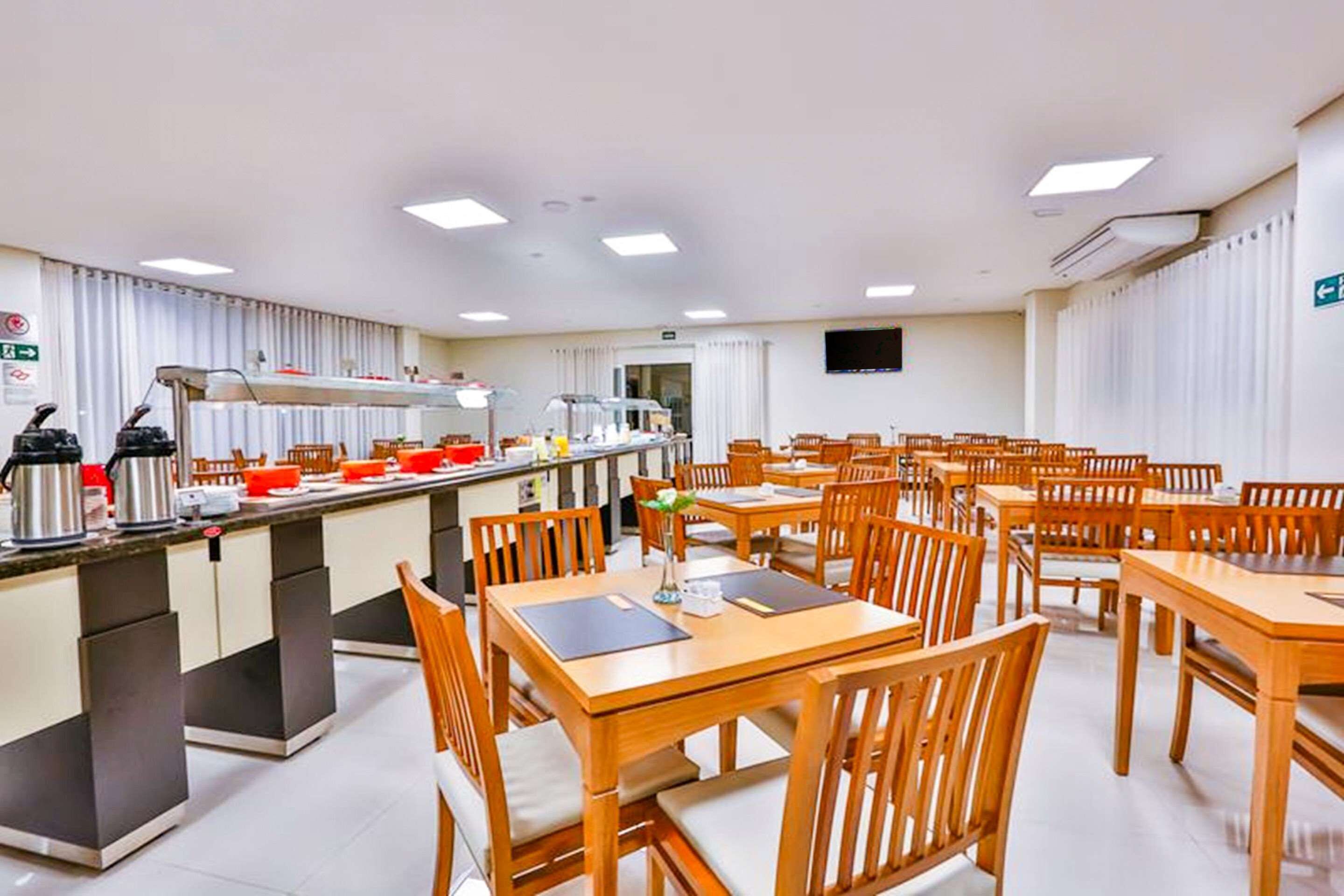A hotel dining area with wooden tables and chairs, a central buffet counter, and a television mounted on the wall.