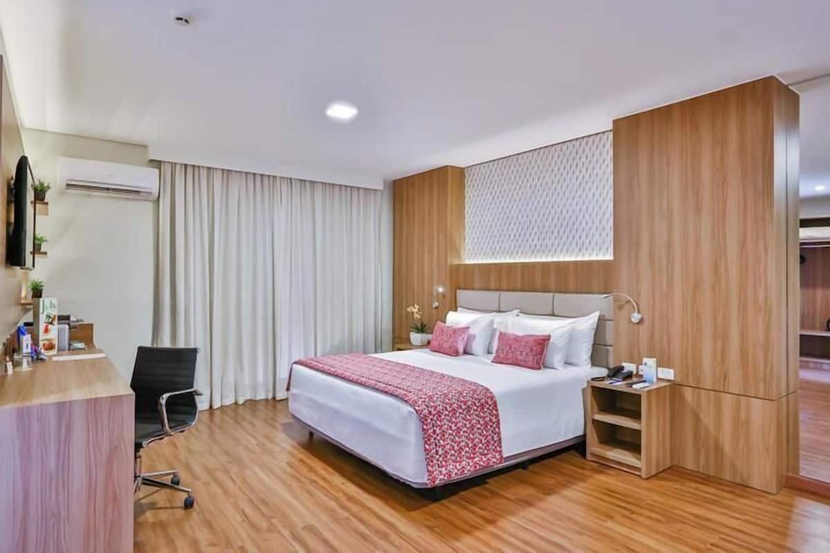 A hotel room with a large bed, a desk, and a chair. There is a window with curtains, a wall-mounted air conditioner, and a wooden panel design on the wall.