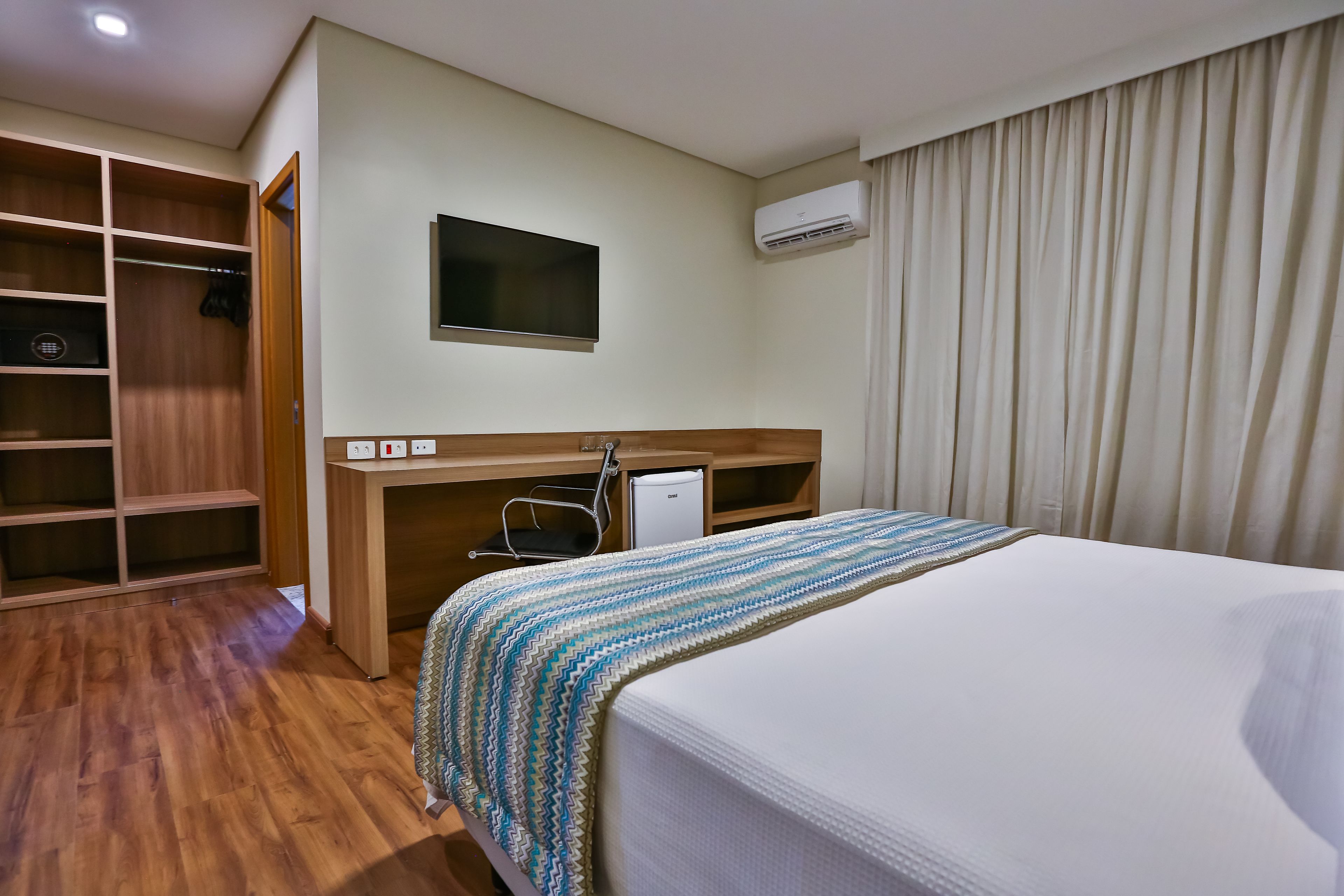 Habitación de hotel con piso de madera, cama con manta a rayas, escritorio con silla, televisor y armario.