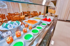 Buffet com diversos alimentos e bebidas, incluindo tigelas com pratos verdes e laranjas, e bandejas de frutas fatiadas.
