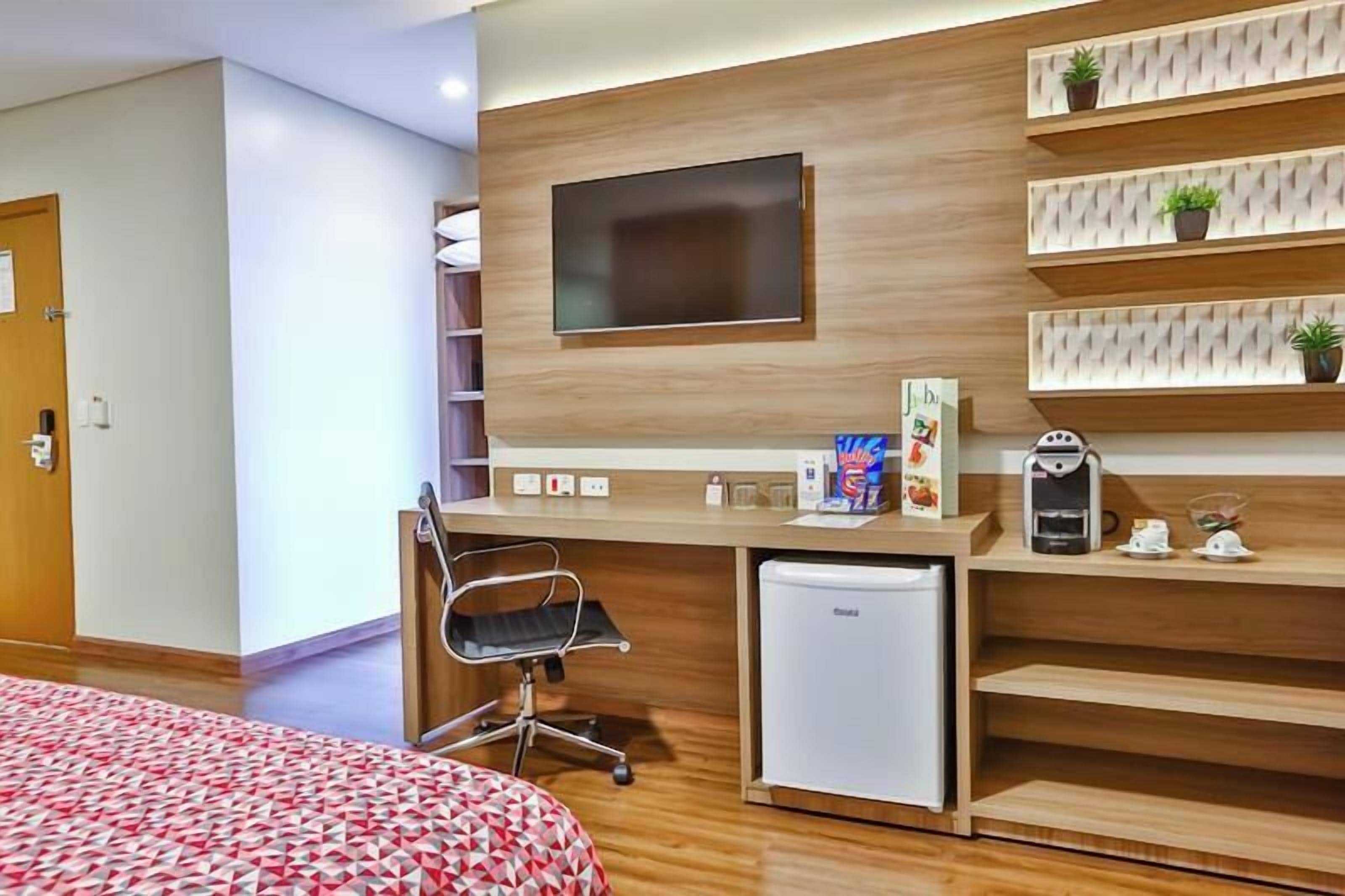 Une chambre d’hôtel moderne dotée d’un bureau en bois, d’un téléviseur à écran plat, d’un mini-réfrigérateur, d’une machine à café et d’un lit recouvert d’une couvre-lit à motifs.