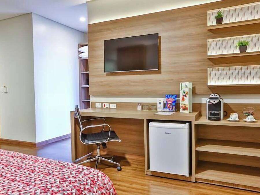 Une chambre d’hôtel moderne dotée d’un bureau en bois, d’un téléviseur à écran plat, d’un mini-réfrigérateur, d’une machine à café et d’un lit recouvert d’une couvre-lit à motifs.