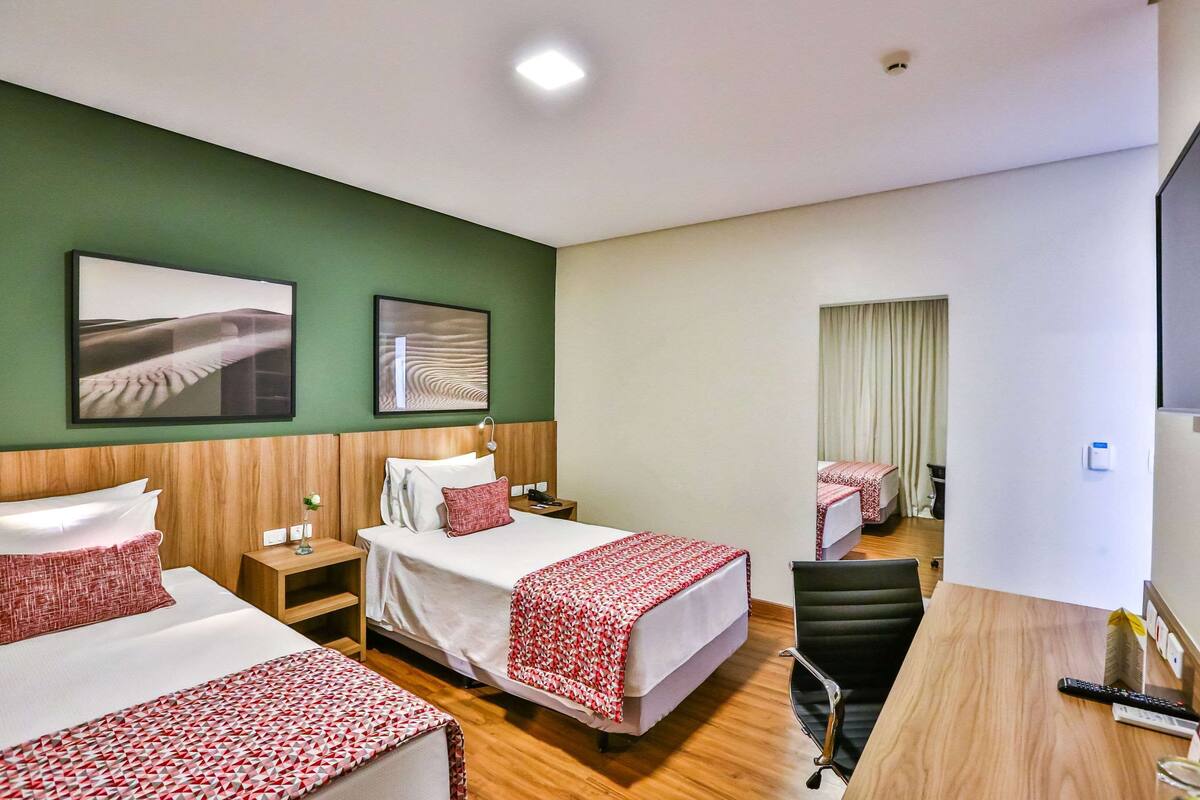 A hotel room with two beds, a desk, and a chair. There are two bedside tables with lamps, a flat-screen TV, and artwork on the walls.