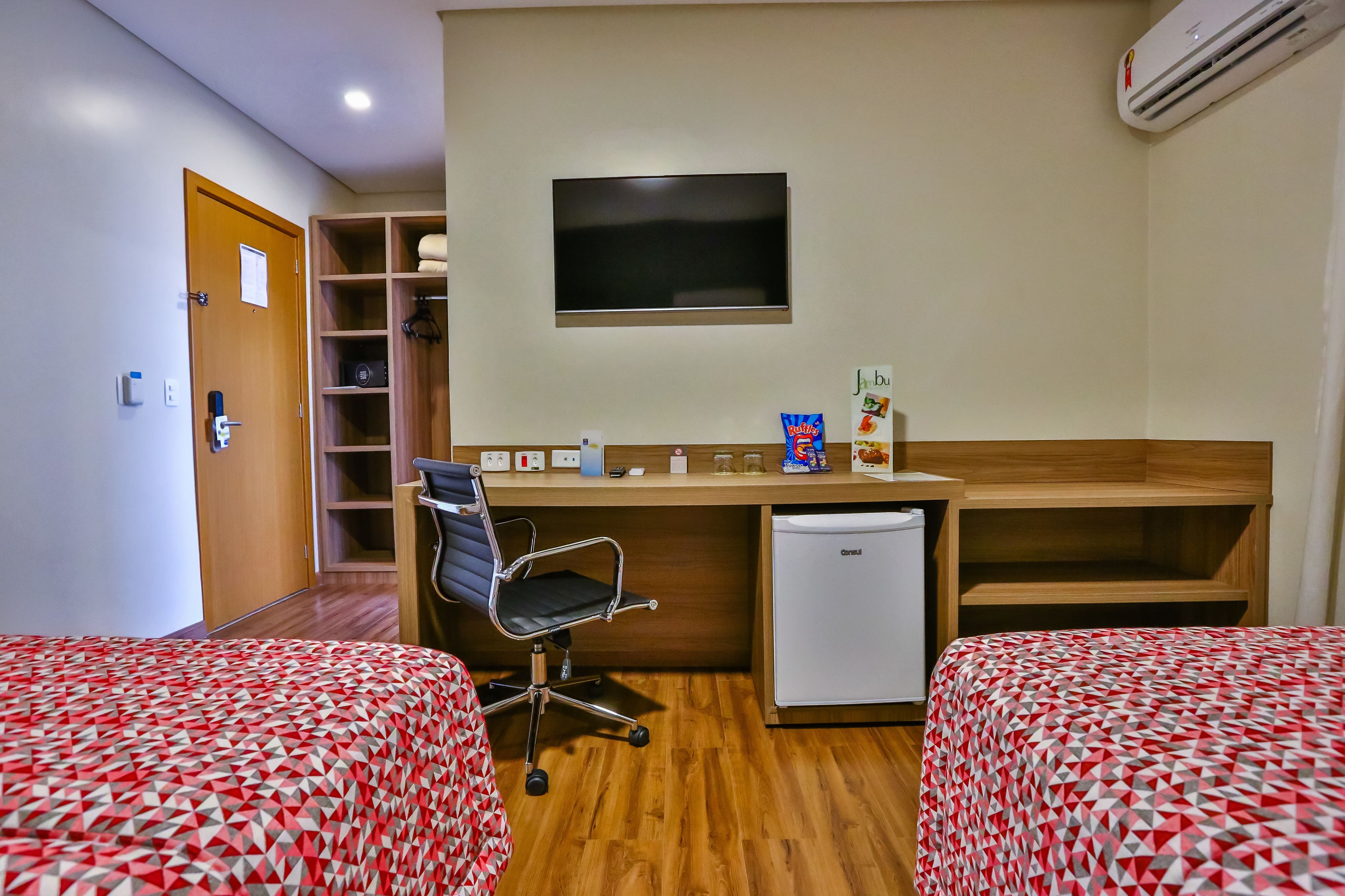A hotel room with two beds, a desk with a chair, a television, a mini-fridge, and a shelf with towels.