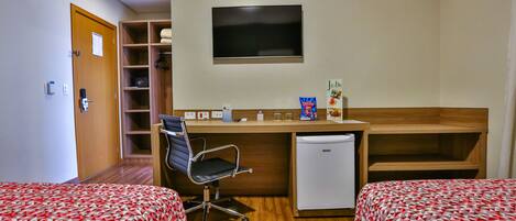 A hotel room with two beds, a desk with a chair, a television, a mini-fridge, and a shelf with towels.