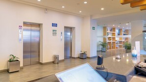 A modern hotel lobby with two elevators, a reception desk, and a decorative area with plants and shelves. - Hotel Misión Puebla Angelopolis (Puebla)
