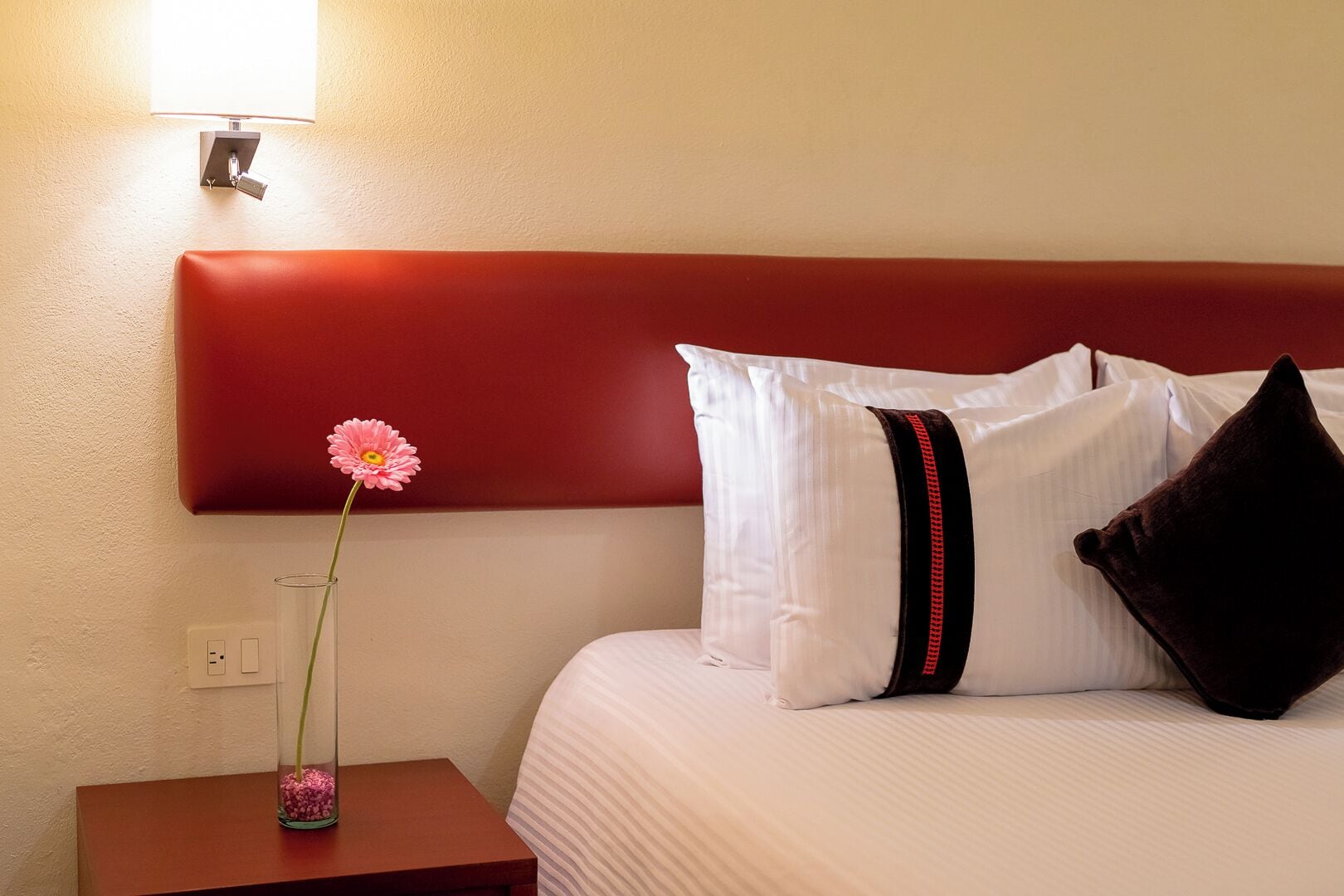 Ein Hotelzimmer mit einem Bett, einem Nachttisch, einer Vase mit einer Blume und einer Wandlampe.