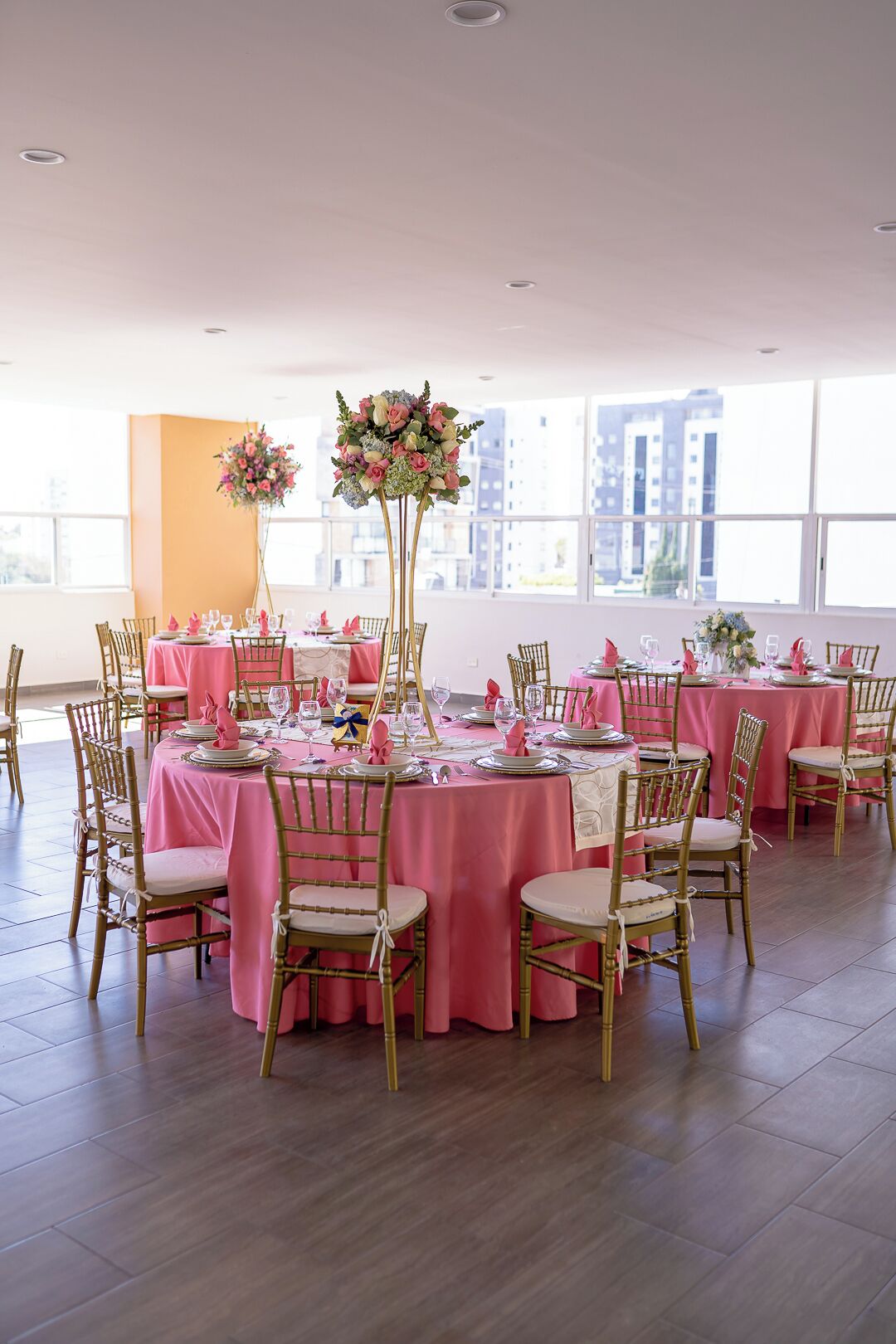 Un salón de banquetes con mesas redondas cubiertas con manteles rosa, sillas doradas y centros de mesa florales.