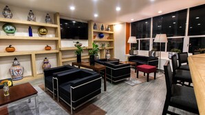 A modern living room with black sofas, wooden shelves with decorative vases, a wooden coffee table, and a large window with a view. - Hotel Misión Puebla Angelopolis (Puebla)