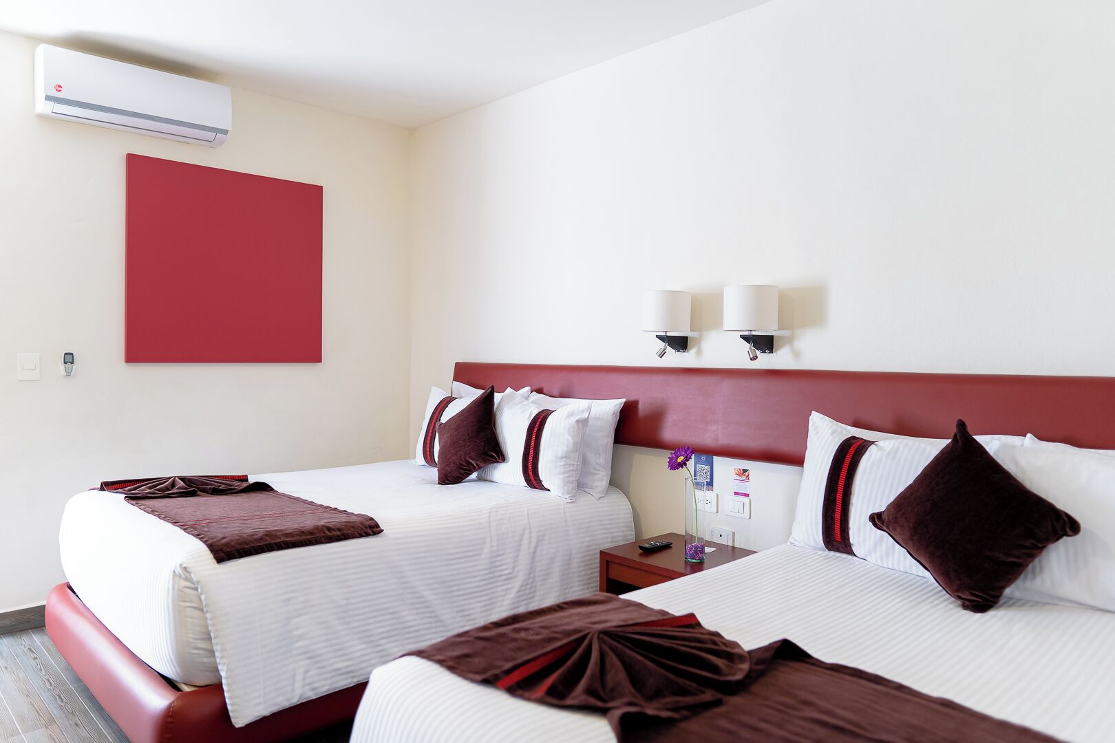 Habitación de hotel con dos camas, una pared decorada en rojo y un aire acondicionado instalado en la pared.