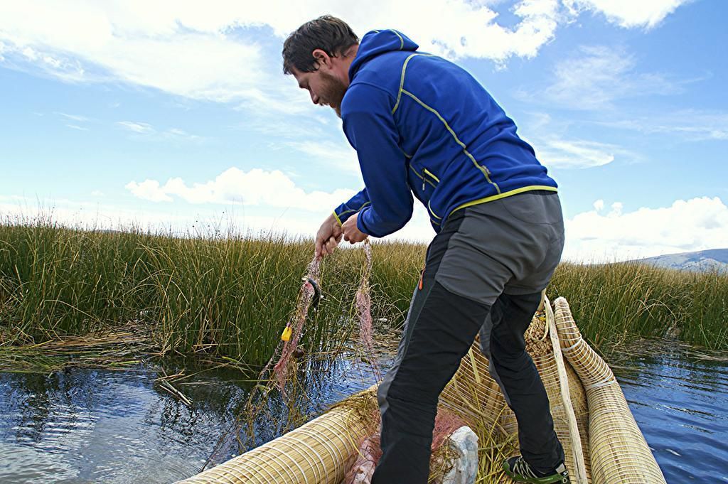 A person in a blue jacket and gray pants standing on a boat in a body of water surrounded by tall grass.