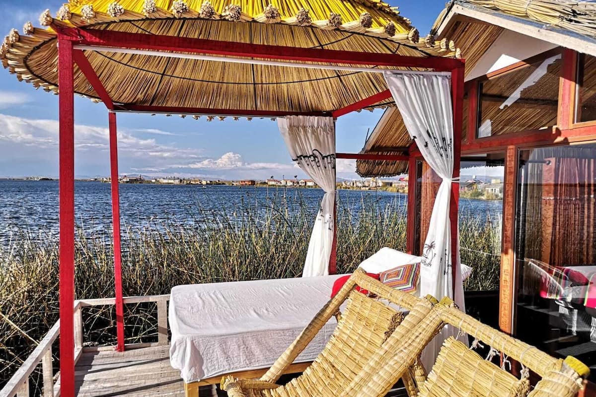 Terraza de madera con una cama y sillones de mimbre, con vista a un cuerpo de agua.