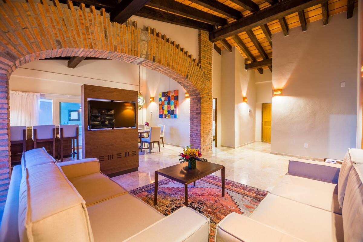 Un salón espacioso con un amplio sofá, una mesa de centro con un arreglo floral y un arco de ladrillo que conduce a otra habitación.