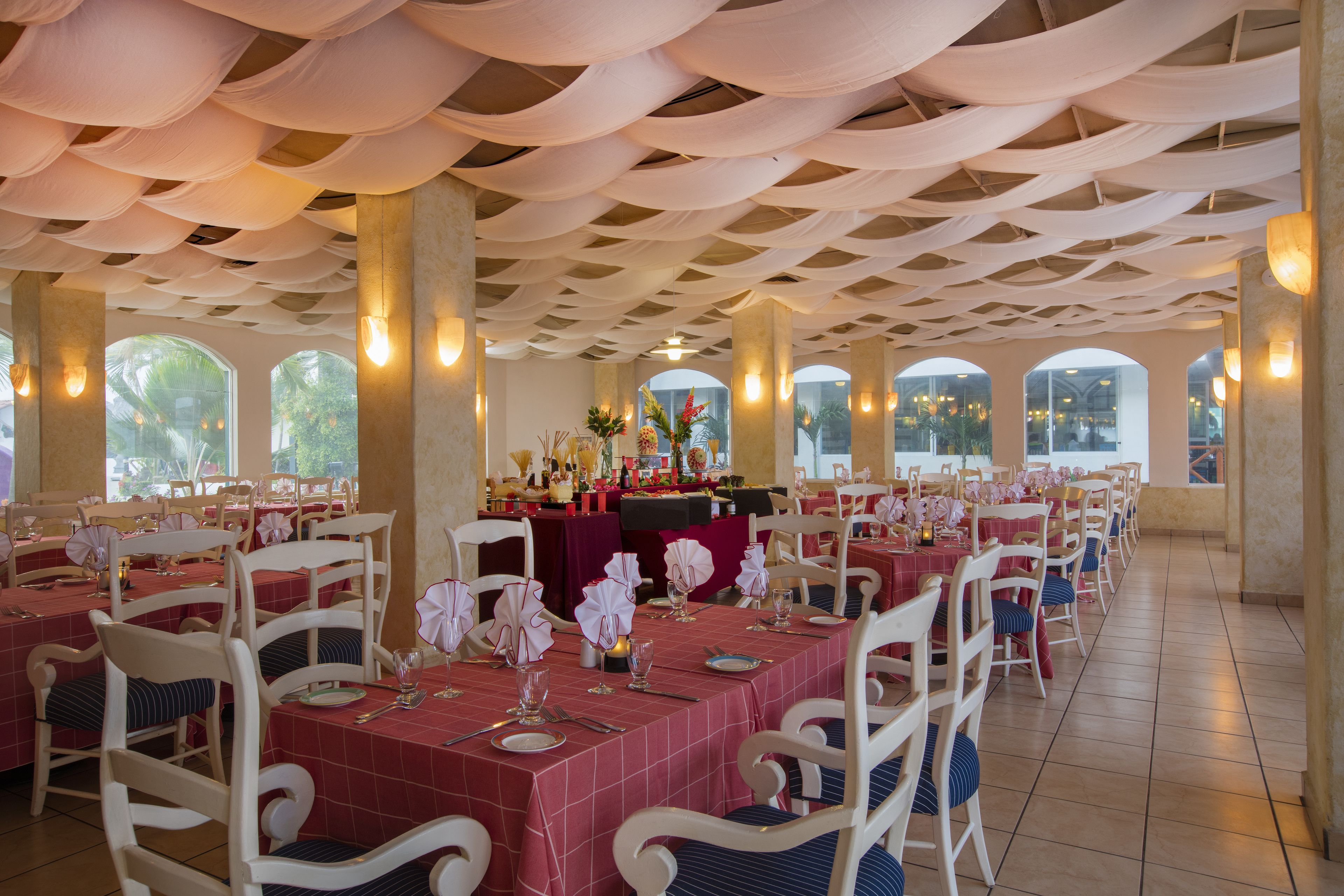 A restaurant with tables set for dining, featuring a ceiling with draped fabric and large windows allowing natural light.