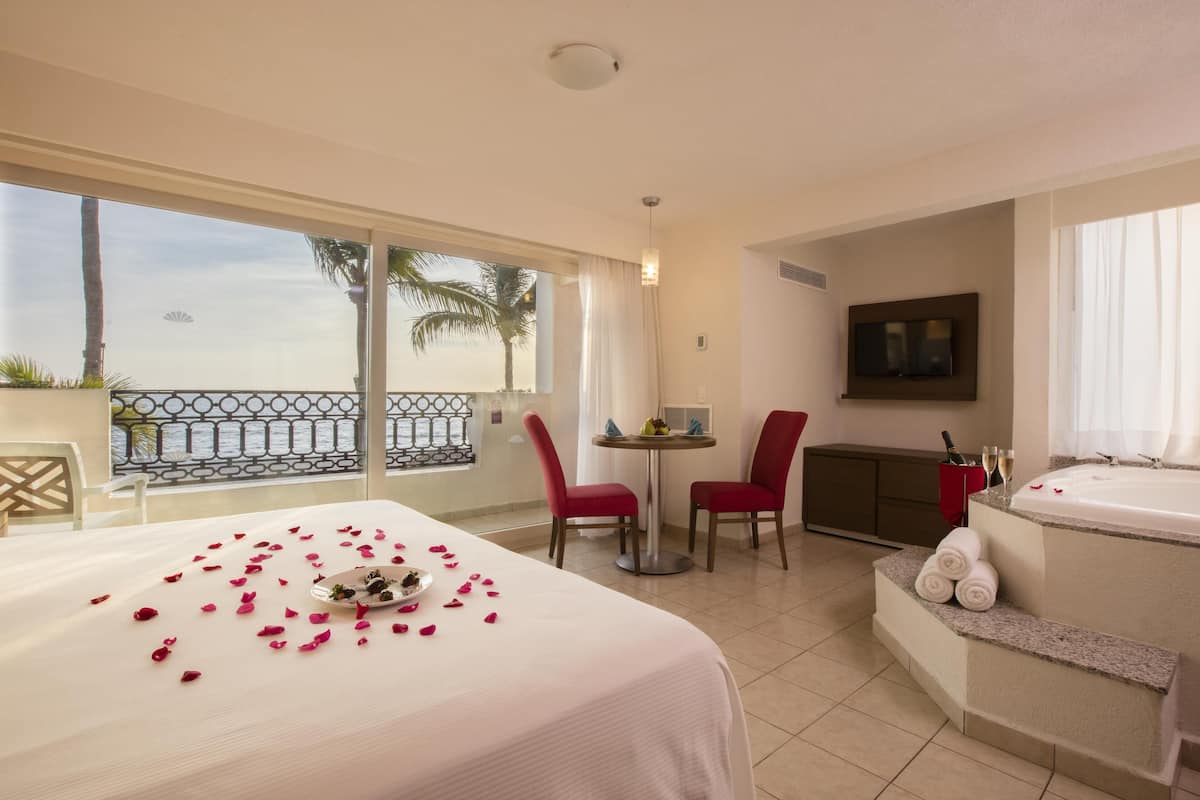 Habitación de hotel con balcón, cama con pétalos de rosa, mesa con sillas, televisor y jacuzzi.