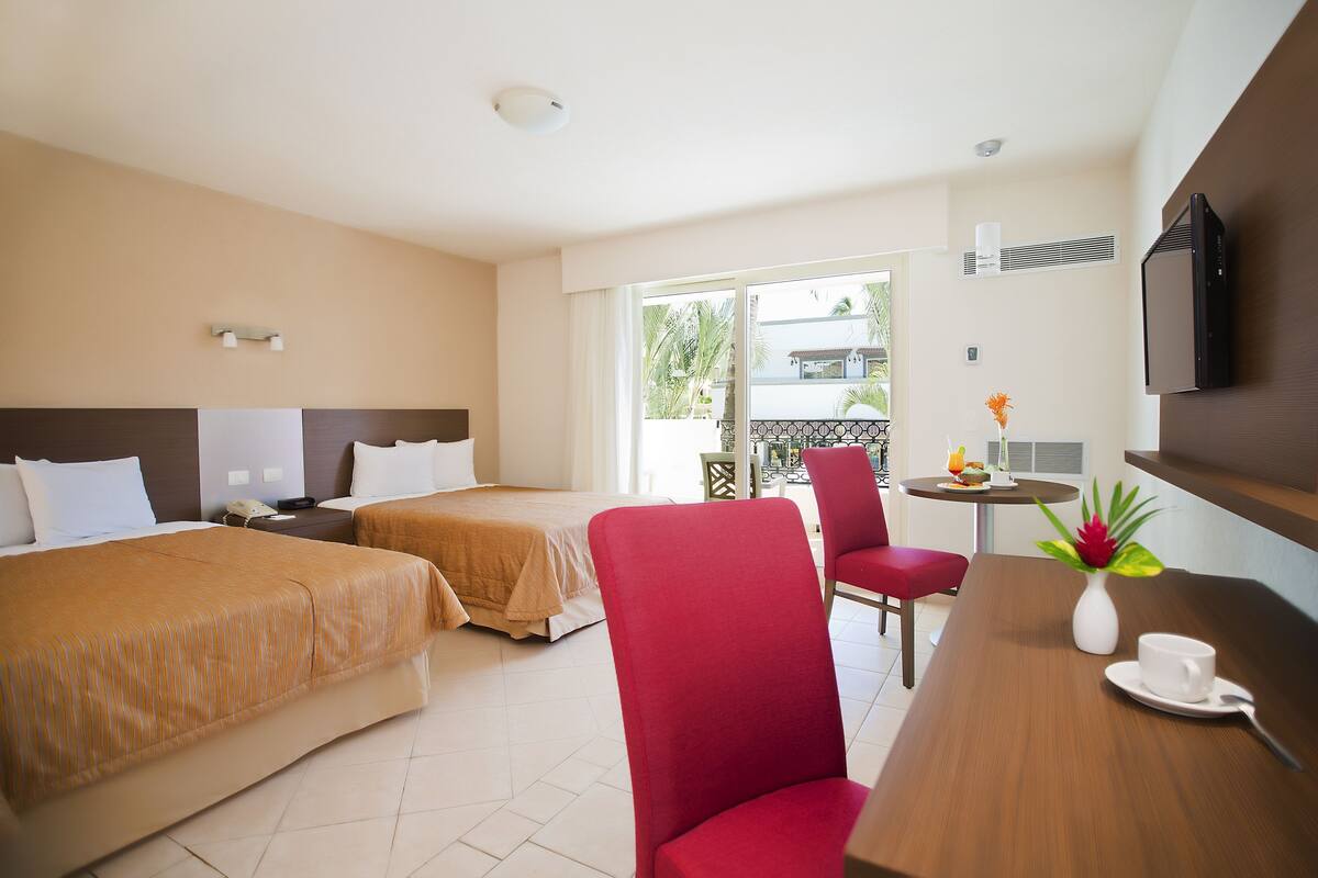 Habitación de hotel con dos camas, un escritorio con silla, una mesita con un florero y una taza, y un televisor montado en la pared.
