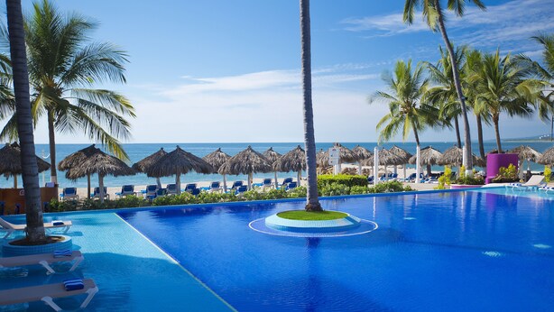 Zona de piscina con sillas de descanso y palmeras, con vista a una playa.