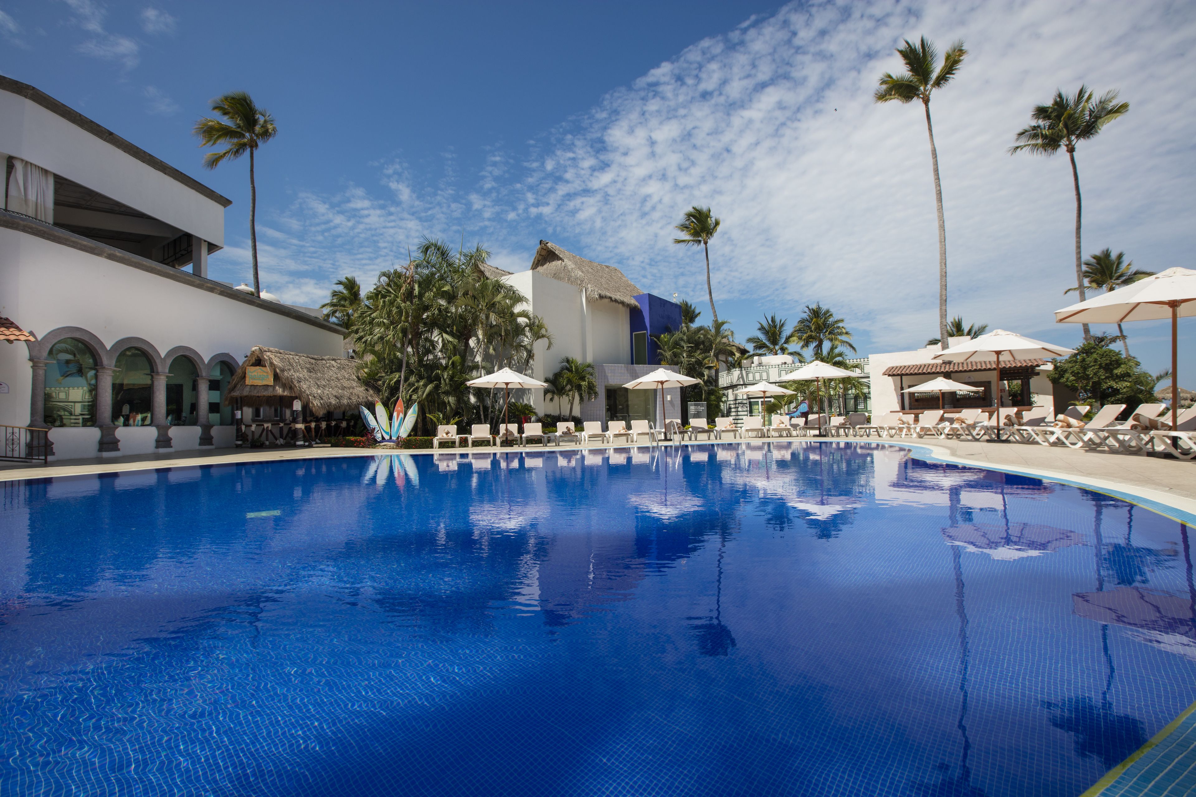 A swimming pool with lounge chairs and umbrellas, surrounded by palm trees and a building with a thatched roof.