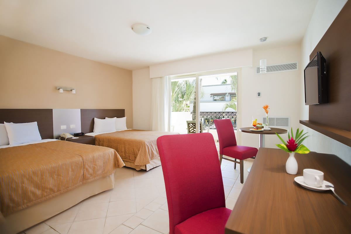 Habitación de hotel con dos camas, una mesa de comedor, una silla roja, una mesita con un jarrón y una taza, y un televisor montado en la pared.