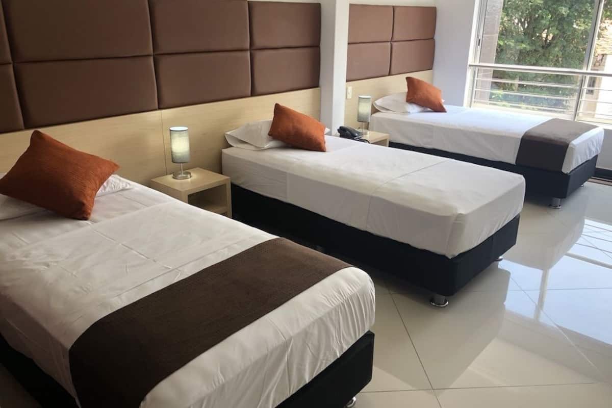 Quarto de hotel com três camas, cada uma com cabeceira marrom, roupa de cama branca e manta marrom. Em cada cama, um criado-mudo com uma luminária e um telefone.