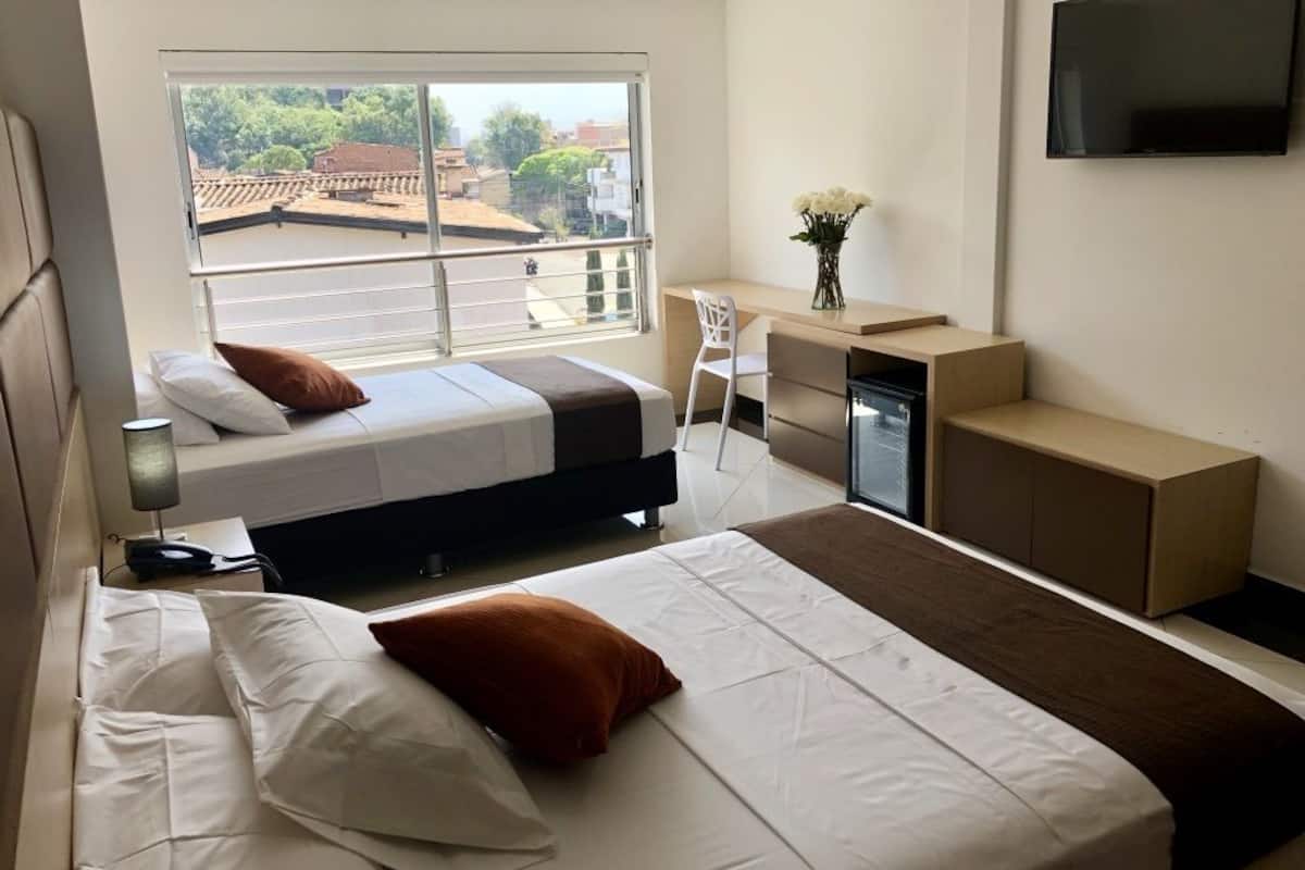 Quarto de hotel com duas camas, uma escrivaninha, televisão e uma janela grande com vista para edifícios e árvores.