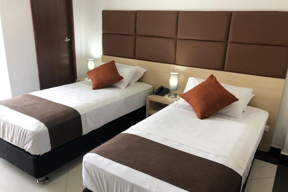 Duas camas de casal com cabeceiras marrons, roupa de cama branca e almofadas decorativas marrons. Cada cama tem uma mesa de cabeceira com uma luminária e um telefone.