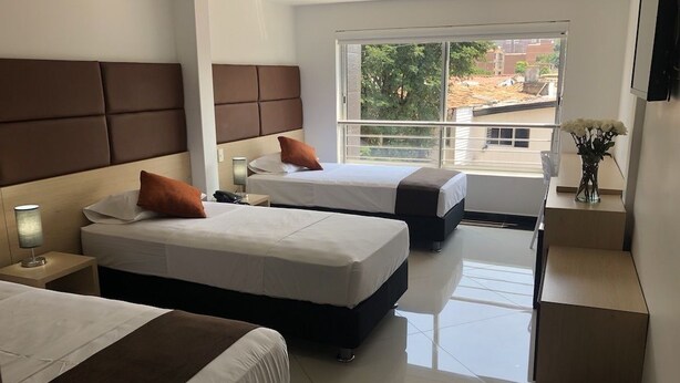 A hotel room with two beds, a small desk, a chair, a nightstand with a lamp, a vase with flowers, and a large window with a view of the outside. - Hotel CSI Medellín (Medellín)