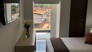 A bedroom with a bed, a nightstand, a vase with flowers, a door, and a view of buildings through a window. - Hotel CSI Medellín (Medellín)