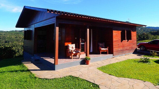 A wooden house with a covered porch, a car parked nearby, and a view of the surrounding greenery. - Residencial Serra Grande Gramado (Gramado)