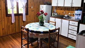 A wooden cabin interior with a dining table, chairs, a kitchen area, and a window with curtains. - Residencial Serra Grande Gramado (Gramado)