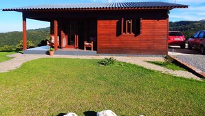 A wooden cabin with a covered porch, a car parked outside, and a view of the surrounding hills. - Residencial Serra Grande Gramado (Gramado)