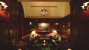A dimly lit lounge area with leather armchairs, a central sofa, a chandelier, and a wall-mounted painting.