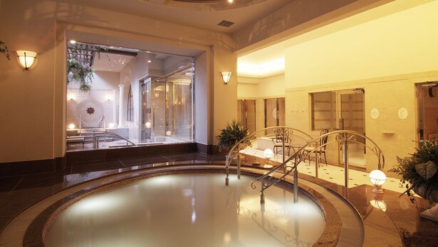 A spacious indoor swimming pool with a glass wall leading to a spa area, surrounded by a well-lit lobby with seating and a staircase.