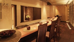 A modern dining area with a long table, chairs, and a central island with a bowl of fruit.