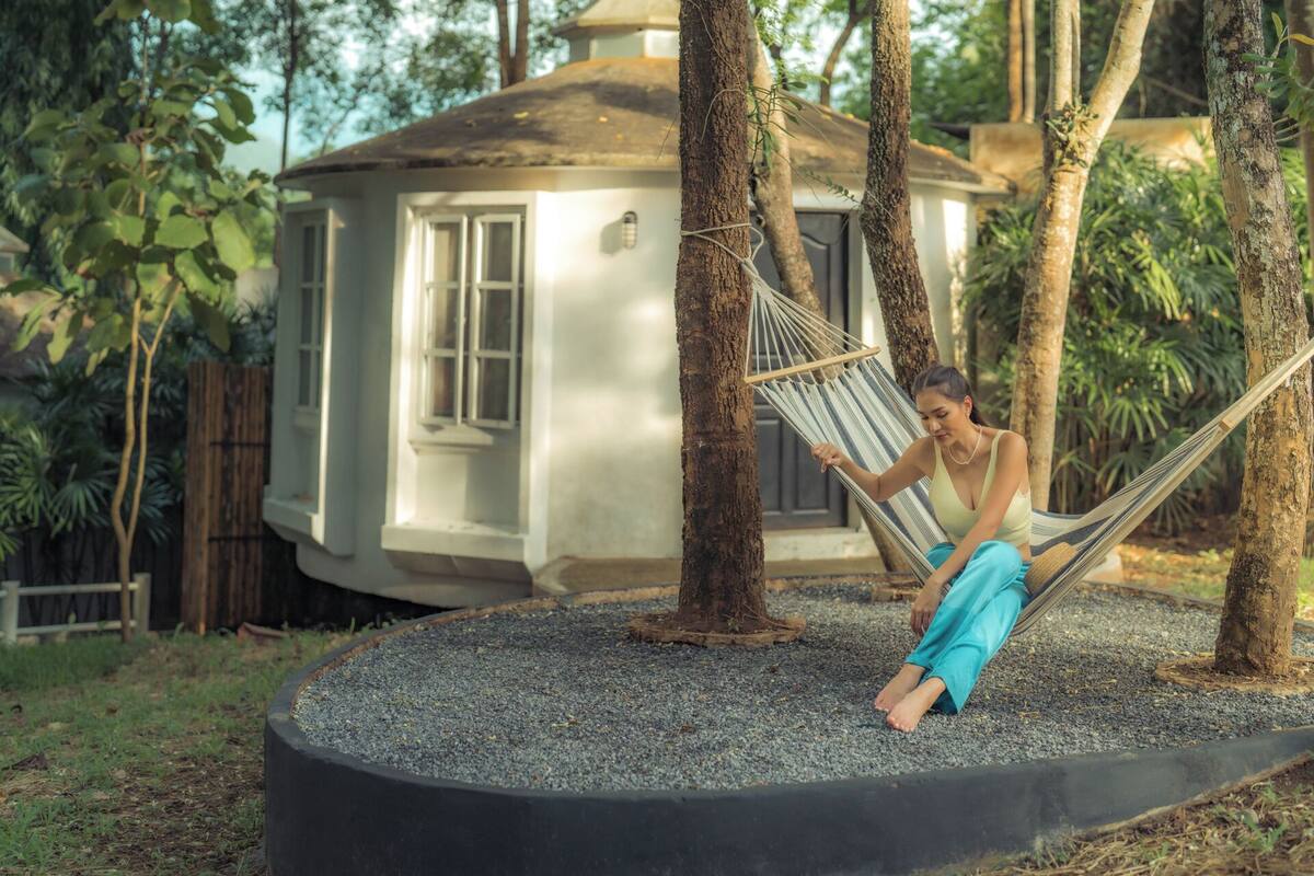 A hammock tied between trees in a garden area with a house in the background.