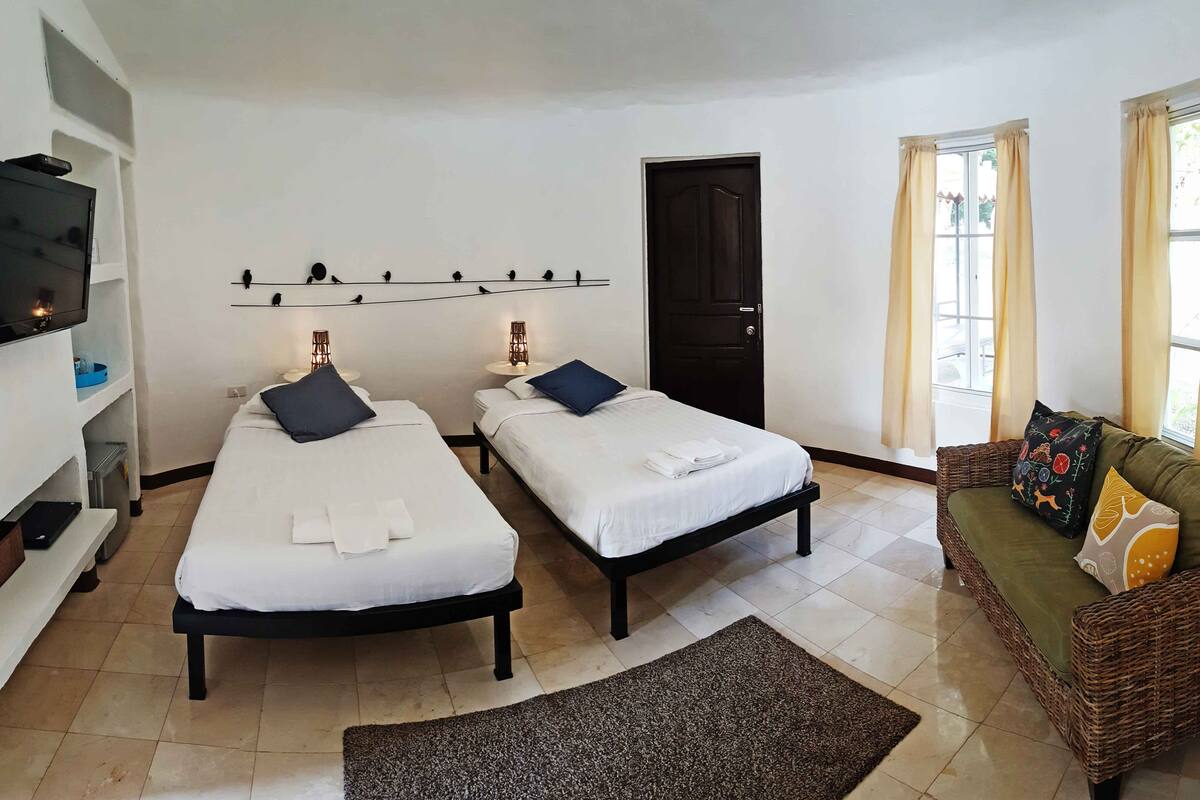 A hotel room with two single beds, a sofa, a TV, and a wall-mounted shelf with decorative items.