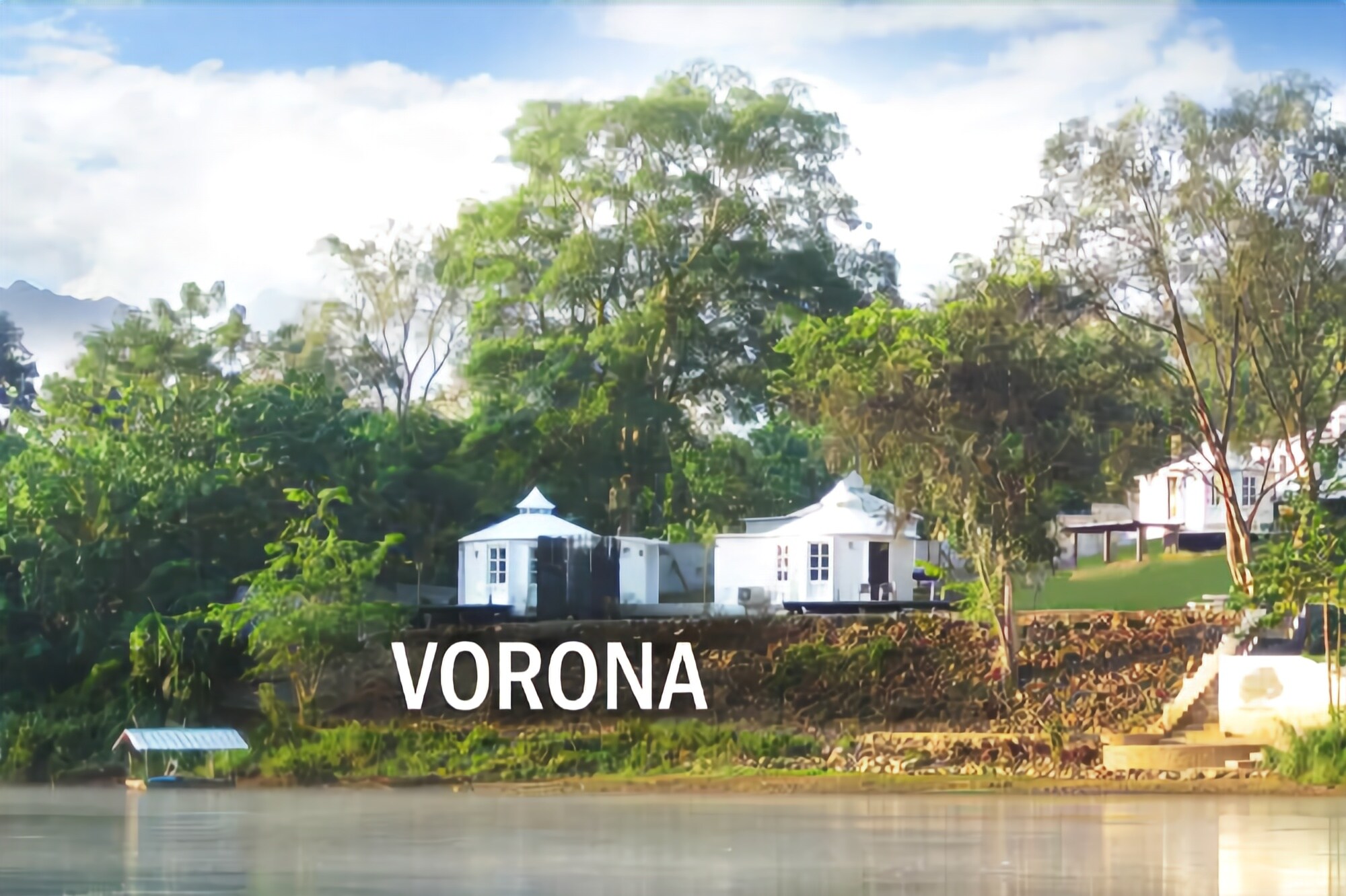 Una propiedad junto al lago con dos edificios blancos, rodeada de vegetación exuberante y un cielo despejado.