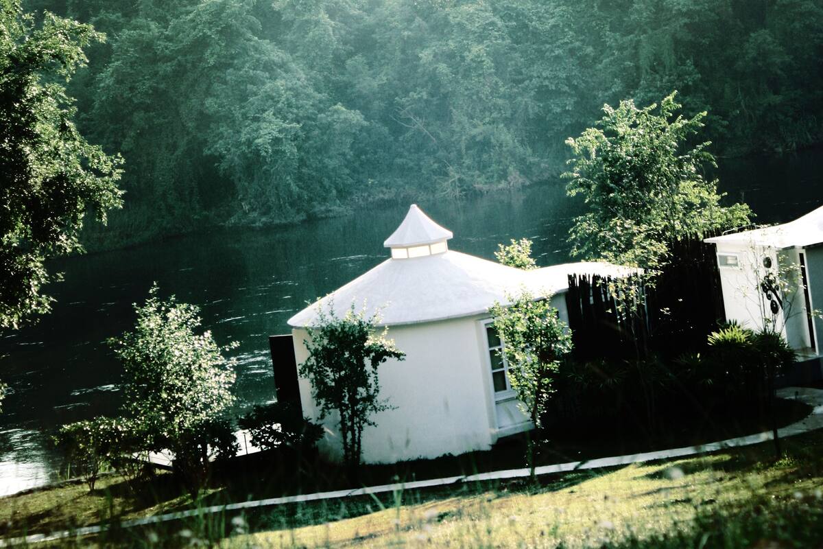 A white building with a conical roof near a body of water, surrounded by dense forest.