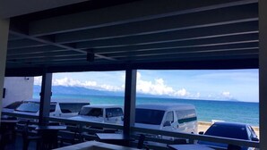 A covered outdoor seating area with tables and chairs, overlooking a beach with parked vehicles and a clear view of the ocean and distant mountains. - Playa Azul Baler (Baler)