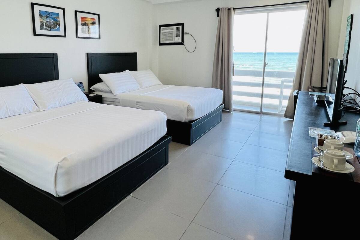 A hotel room with two beds, a desk with a TV, and a view of the sea.