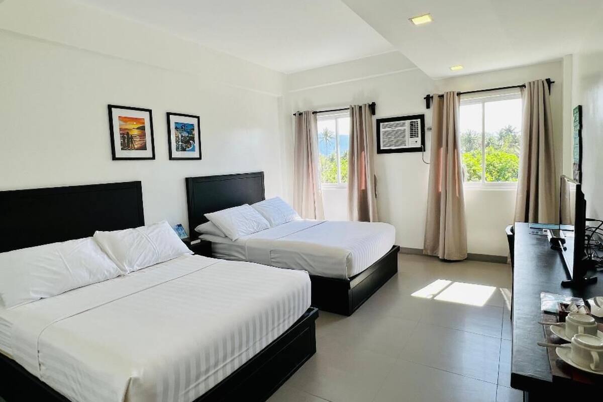 A hotel room with two beds, a desk with a chair, a window with curtains, and a wall-mounted air conditioner.
