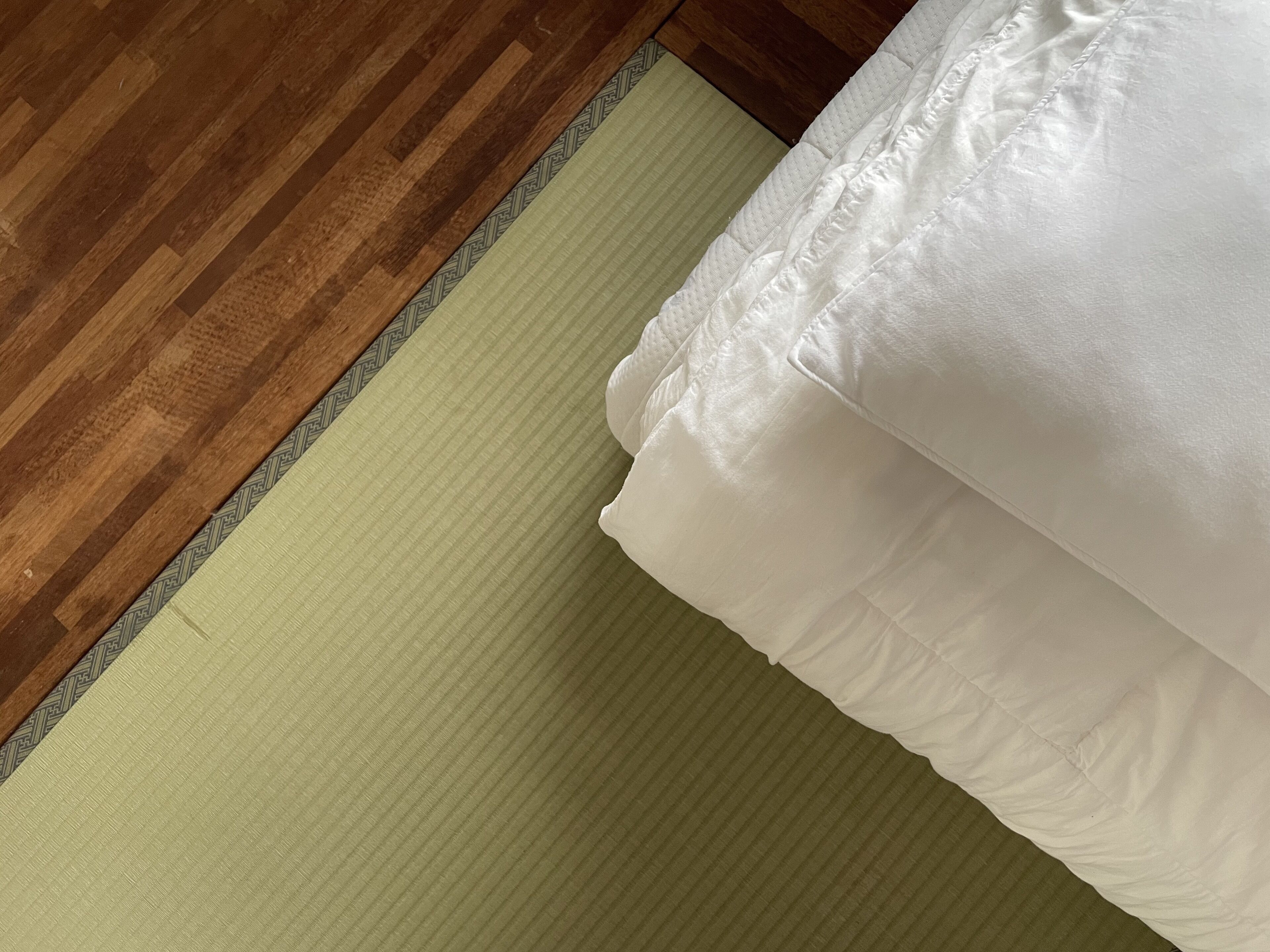 A wooden floor next to a green mat and a white bed with a pillow.