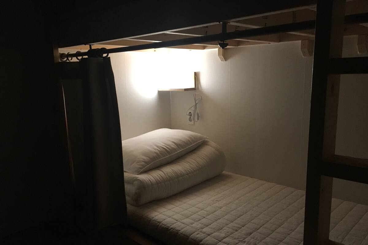 A small, dimly lit room with a single bed, a wall socket, and a window with a curtain.