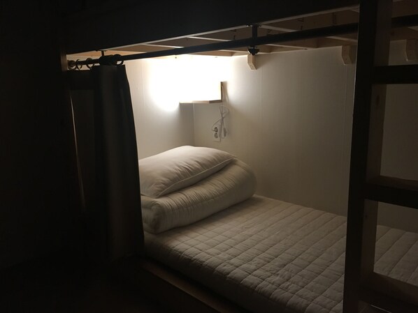 Una habitación pequeña y tenuemente iluminada, con una cama individual, un enchufe de pared y una ventana con cortina.