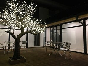 Un espacio al aire libre con mesas y sillas, rodeado de luces que iluminan un árbol.
