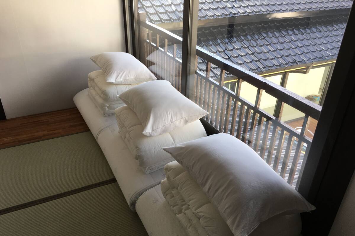 A room with tatami mats, a low wooden bed with white pillows, and a view of traditional tiled roofs through a window.