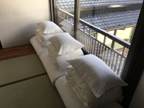 Ein Zimmer mit Tatami-Matten, einem niedrigen hölzernen Bett mit weißen Kissen und Blick auf traditionelle Ziegeldächer durch ein Fenster.